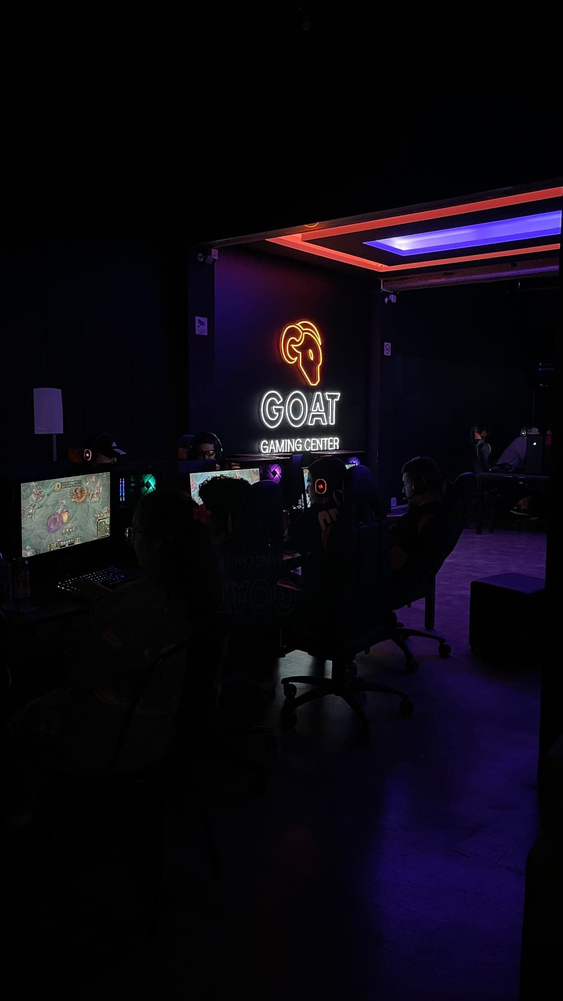 Salão da GOAT Savassi à meia-luz com PCs gamer e o logo GOAT na parede